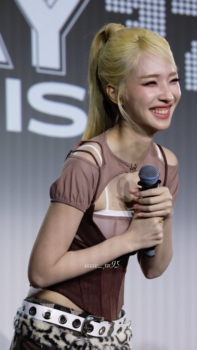 A blonde woman with her hair in a ponytail smiles while holding a microphone, wearing a sheer top and leopard print pants.