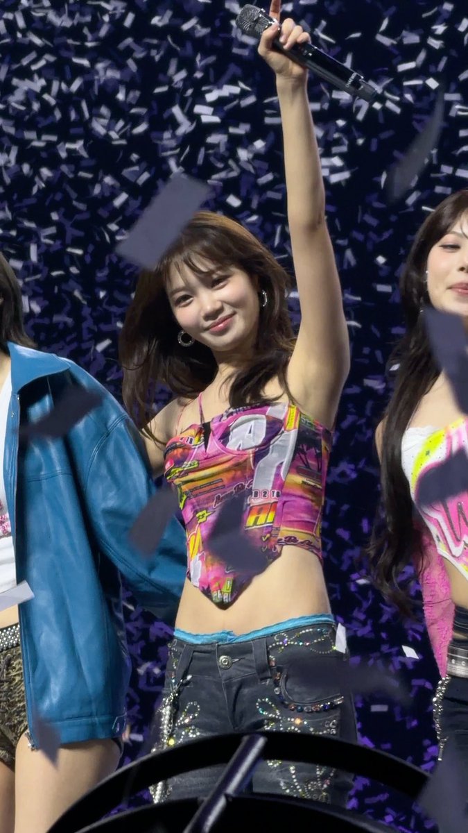 A young woman in a colorful patterned crop top smiles and holds a microphone aloft amid falling confetti at a concert.