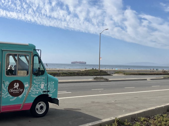 A teal 'Churned Cup' ice cream and coffee truck parked along a coastal road, with a large cargo ship visible on the ocean in the background under a partly cloudy sky.