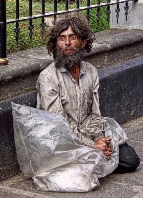 A man with long unkempt hair and a beard, wearing dirty clothes and sitting on a sidewalk with a plastic sheet.