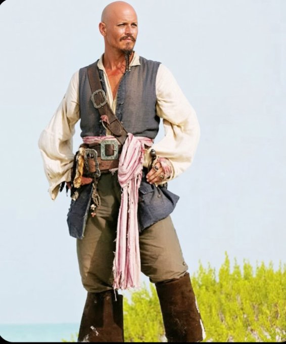 A bald man with a braided goatee stands outdoors wearing a pirate-style outfit with a brown vest, white shirt, sash, and a large belt buckle.