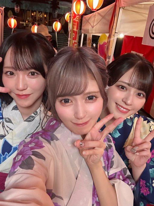 Three young women in floral kimonos pose together for a selfie, making peace signs, with red paper lanterns and festival stalls in the background.