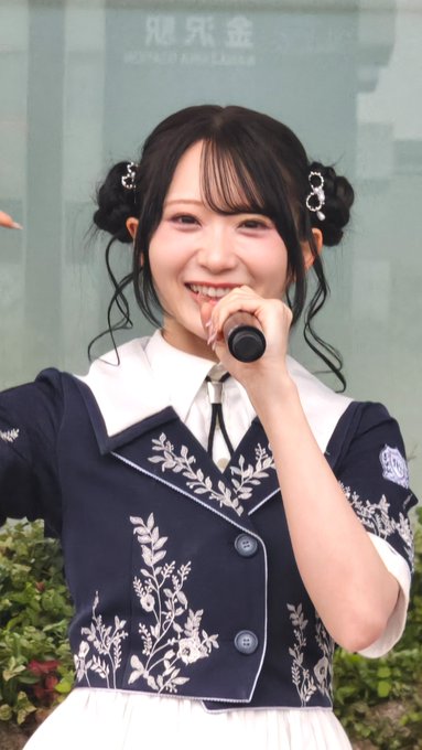 A smiling young woman with her hair in double buns wears a dark blue vest with white floral embroidery over a white collared shirt, holding a microphone.