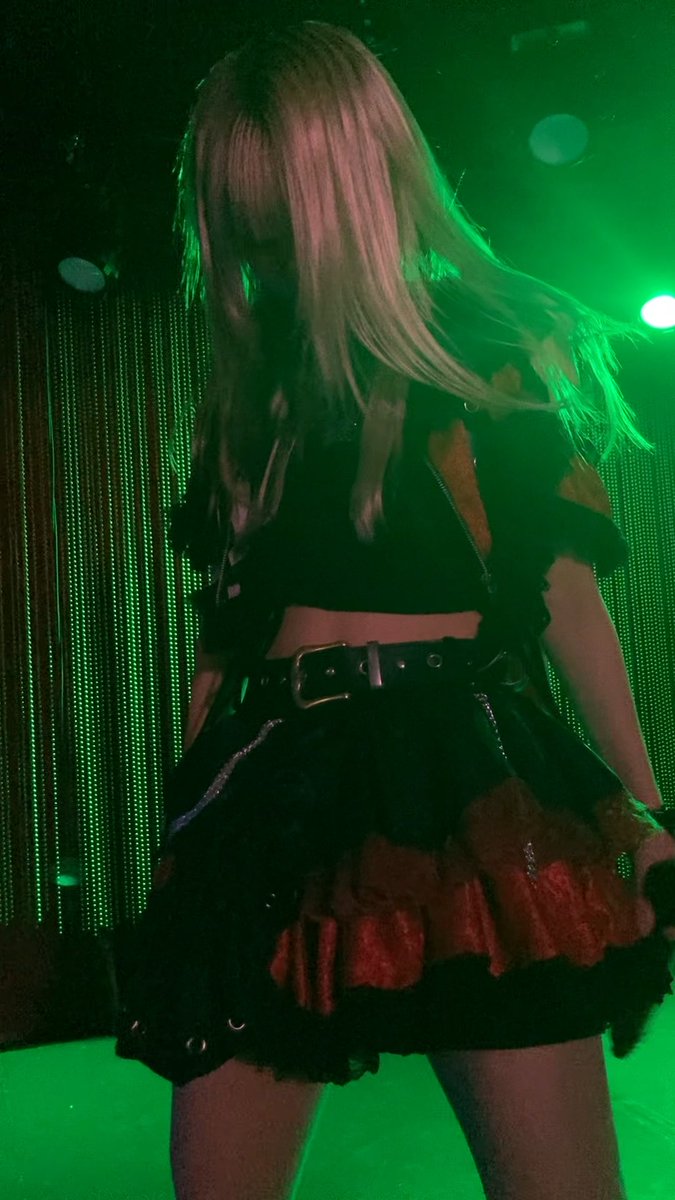 A person with long blonde hair in a dark gothic-style dress and red ruffled skirt stands against a backdrop illuminated by green lighting.