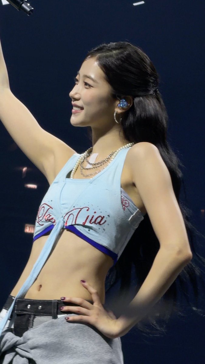 A smiling young woman with long dark hair performs on stage, wearing a light blue crop top with red lettering, layered necklaces, and an in-ear monitor, with one arm raised.