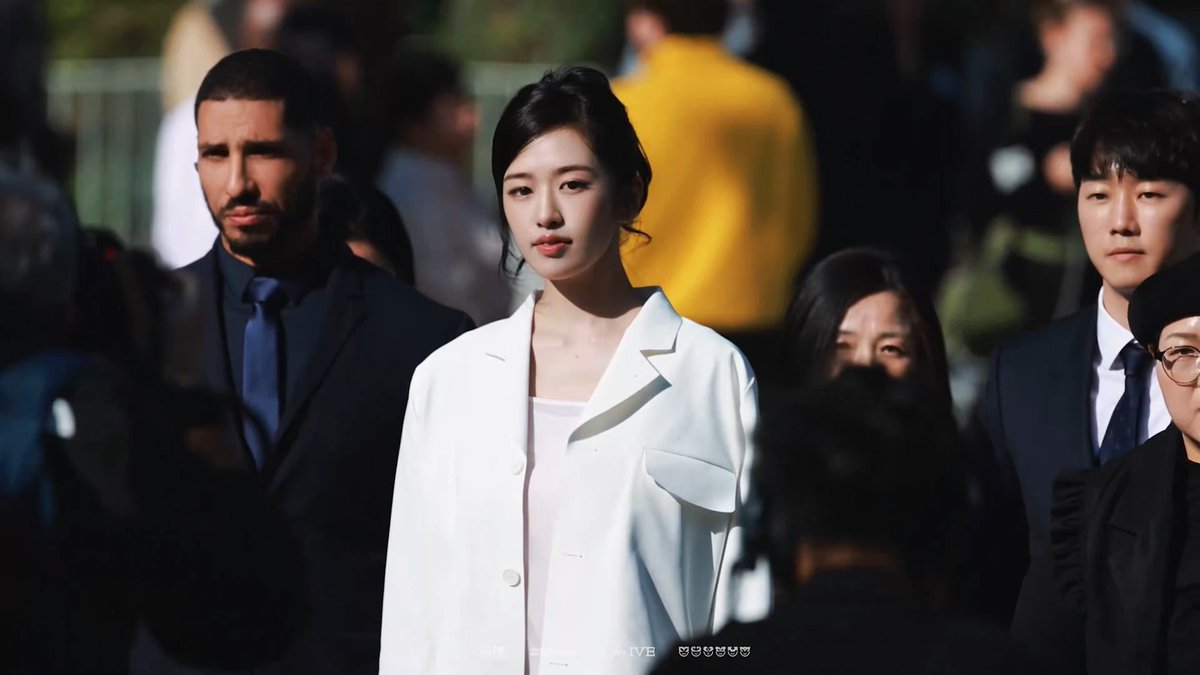 A young woman with dark hair pulled back, wearing a white blazer, stands in a crowd with security personnel, including a man in a dark suit, visible around her.