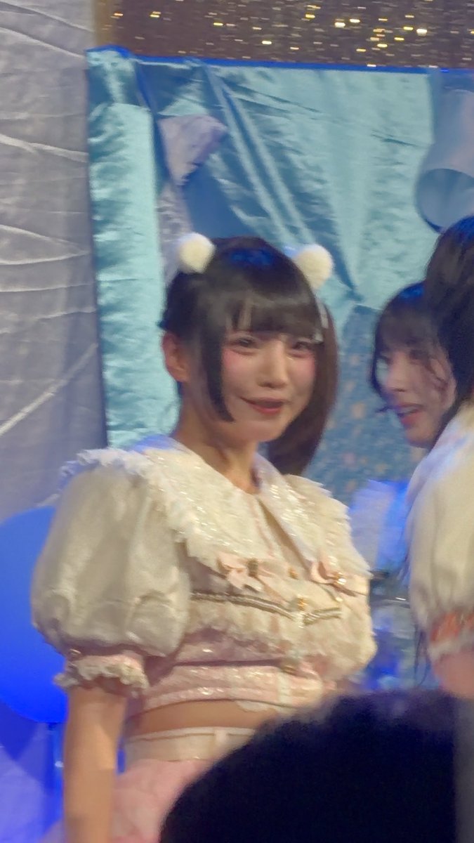 A young woman with white fluffy hair accessories wearing a frilly white and pink outfit, photographed at what appears to be a concert or event.