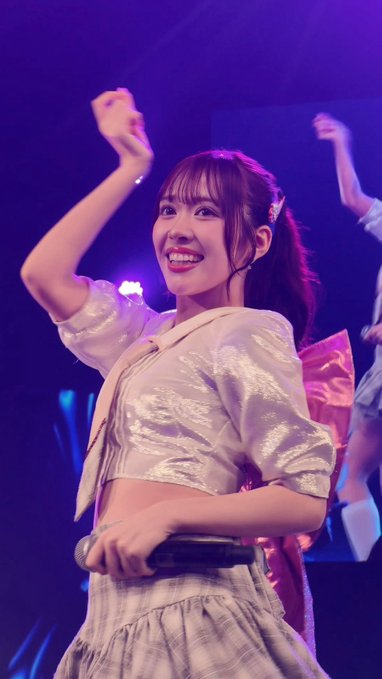 A young woman with a ponytail smiles brightly while performing on stage, holding a microphone and wearing a sparkly white top with a pink bow under purple concert lighting.