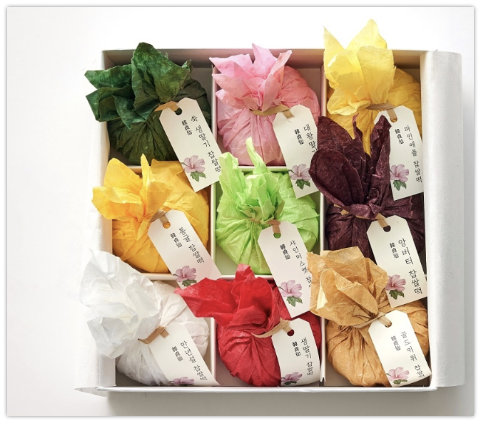 A white gift box containing nine colorful tissue-wrapped bundles in a 3x3 arrangement, each tied with a string and tagged with a label featuring Korean text and a small pink flower illustration.