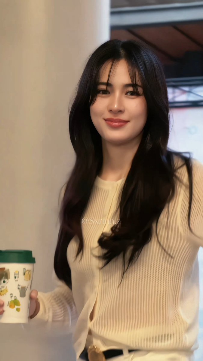 A young woman with long dark wavy hair wearing a cream-colored knit sweater, holding a decorated cup with a green lid, with a watermark '@Sxrinz' visible.
