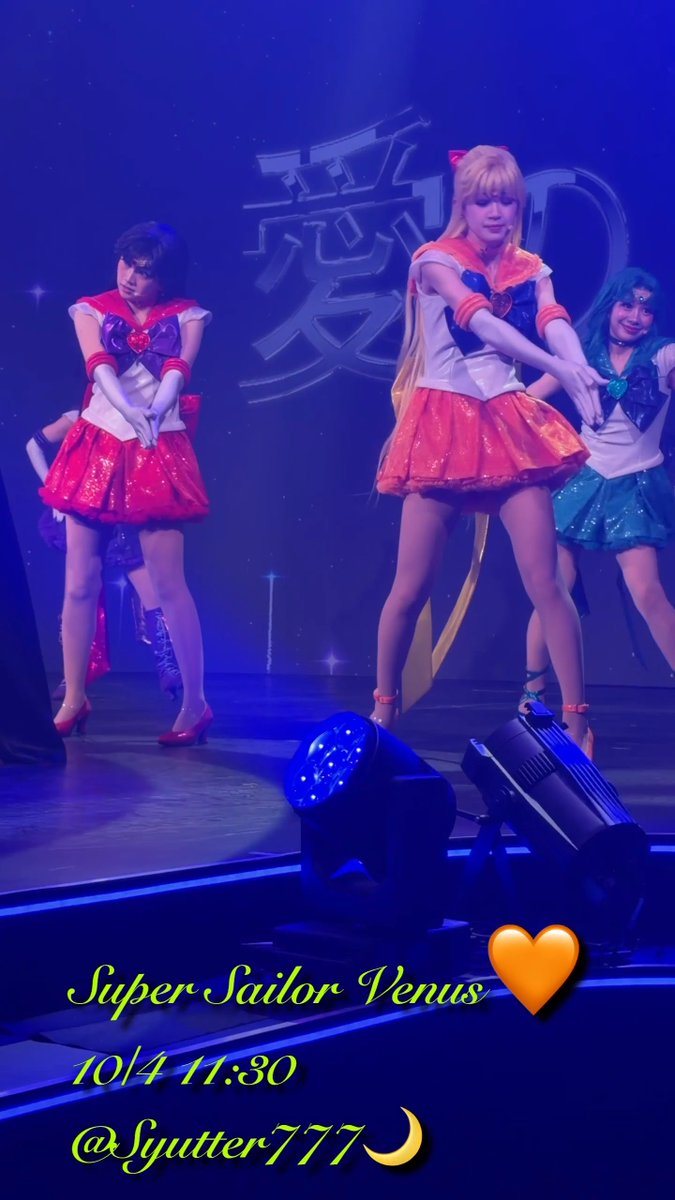 Three actresses strike synchronized poses on a blue-lit stage in sailor-style costumes featuring red and teal skirts, with Japanese characters visible on the backdrop. The image is captioned "Super Sailor Venus" with the date "10/4 11:30" and tagged "@Lyutter777," highlighting a performance from the Sailor Moon Shining Theater, featuring Minako (Venus).