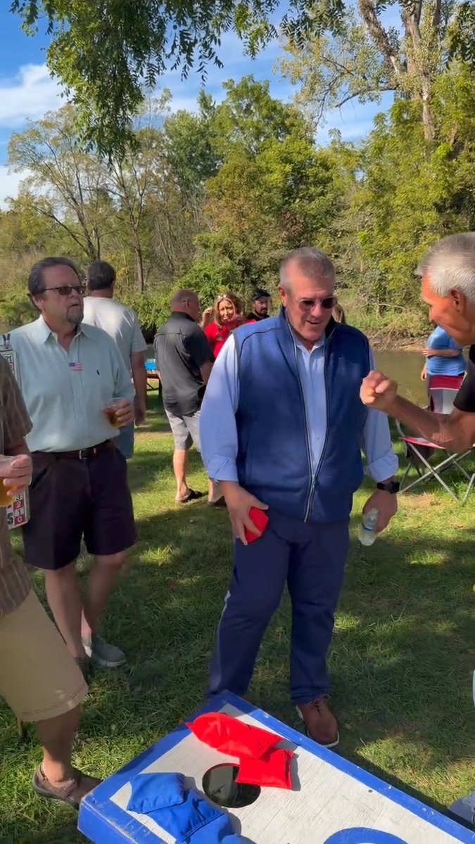 A group of people, including a man in a blue vest holding a red beanbag, stand outdoors near a cornhole board at a grassy gathering by the water.