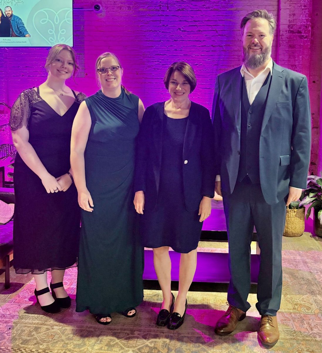 Four adults pose together for a photo in a venue with purple lighting and a brick wall. A display with text is visible in the upper-left corner.