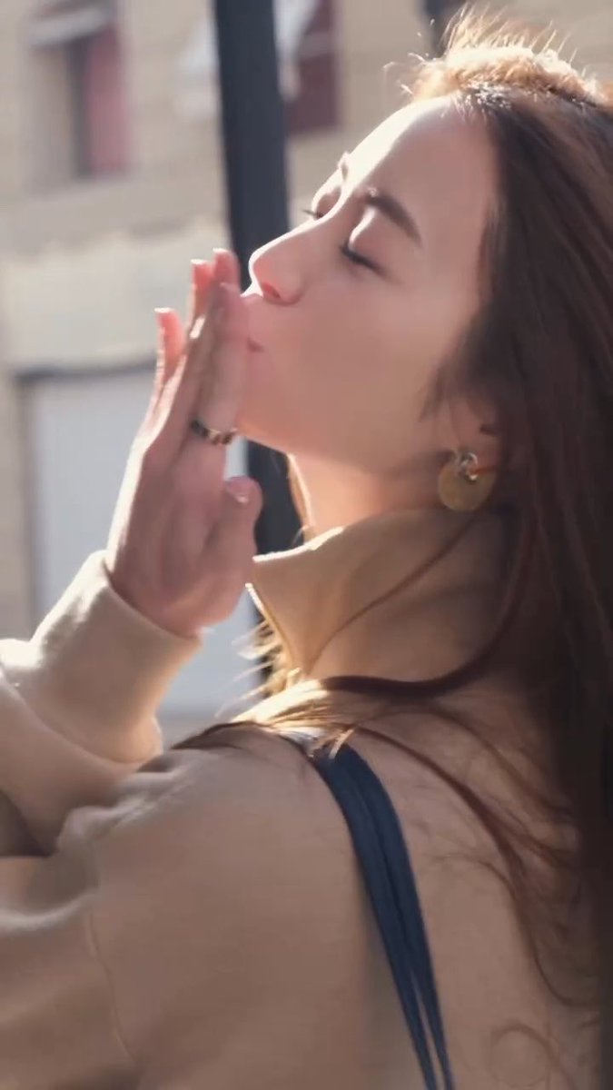 A close-up photograph of a young woman with long brown hair, eyes closed and face tilted up toward the sunlight, wearing a beige turtleneck and gold disc earrings, with her hand gently touching her face.
