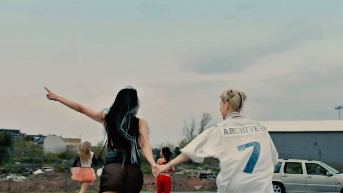Two young people, a woman with long black hair and a blonde man wearing a white shirt with 'ARCHIVE' and the number '7' on the back, hold hands while running towards the horizon in an open outdoor area.