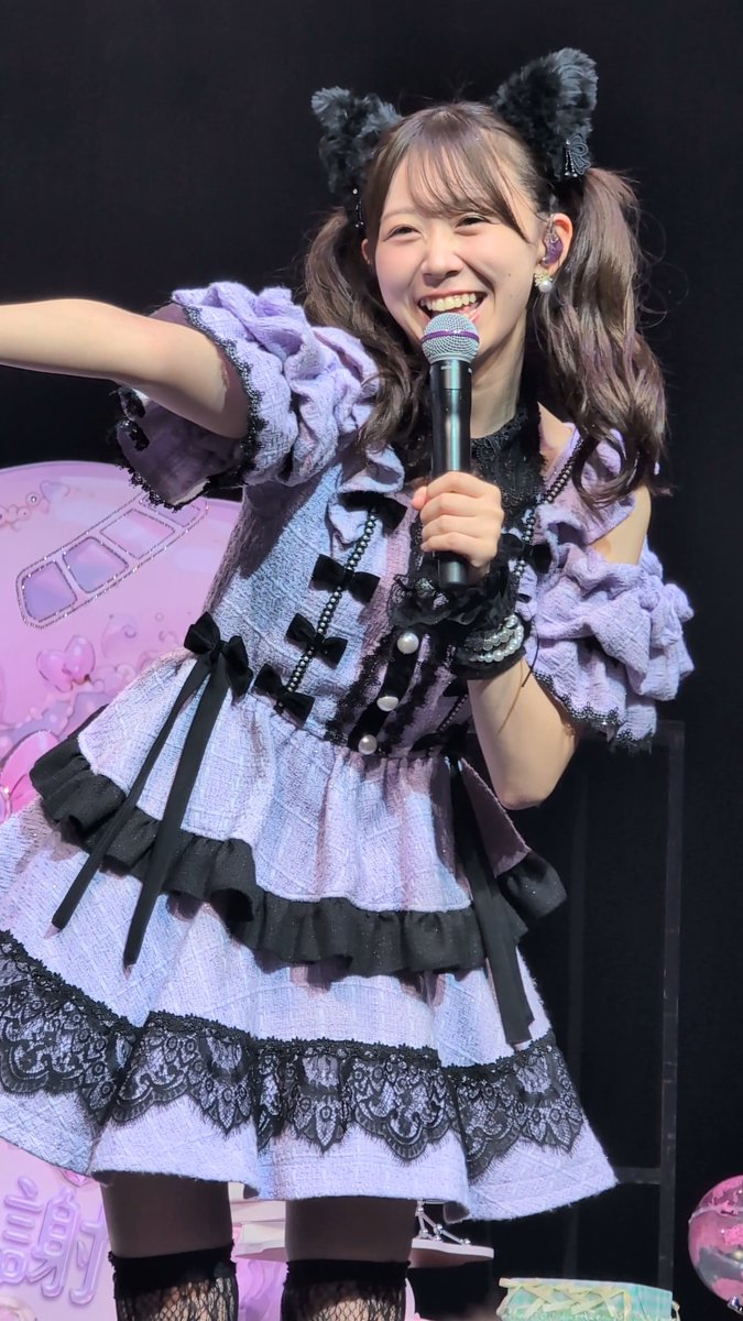 Photo of a smiling young woman performing on stage, wearing a purple and black lolita-style dress with bows, black ribbons, and fluffy cat ear headbands, holding a microphone.