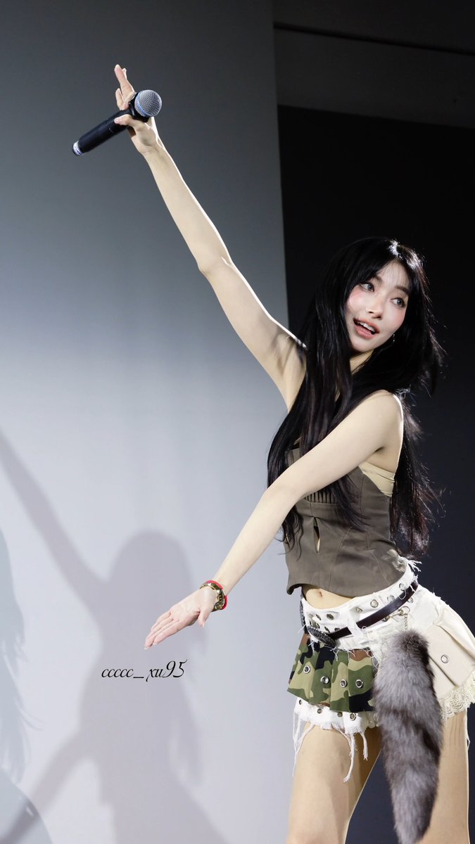 A performer on stage holding a microphone aloft, wearing a tan crop top and a layered skirt with camo and lace details, with a watermark reading 'ccccc_ru95'.