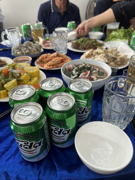 A table is set with various Vietnamese dishes, including seafood, vegetables, and multiple green 'Bia Hanoi' beer cans.