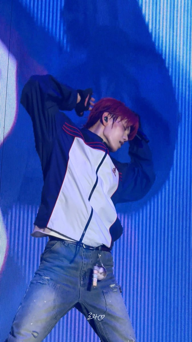 A performer with reddish-brown hair in a white and black track jacket and ripped jeans dancing on a stage with a blue LED background, with a '324∞' watermark.