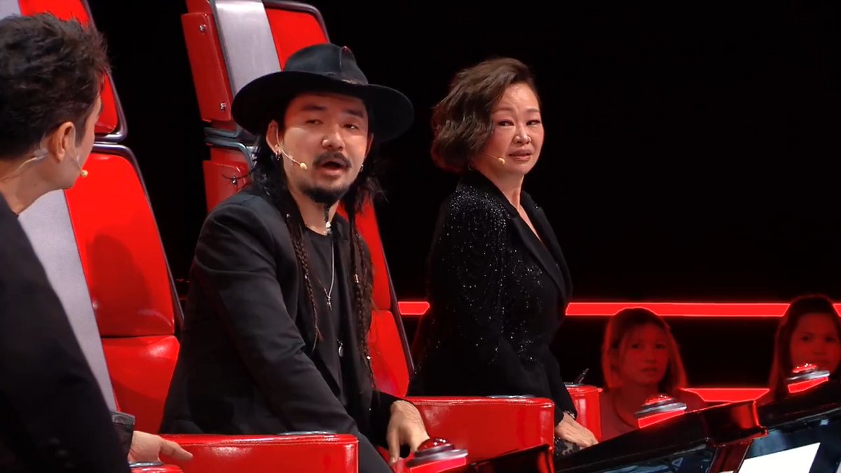 A scene from what appears to be a singing competition show, showing a man in a black hat and jacket seated in a red chair next to a woman in a black sequined blazer.