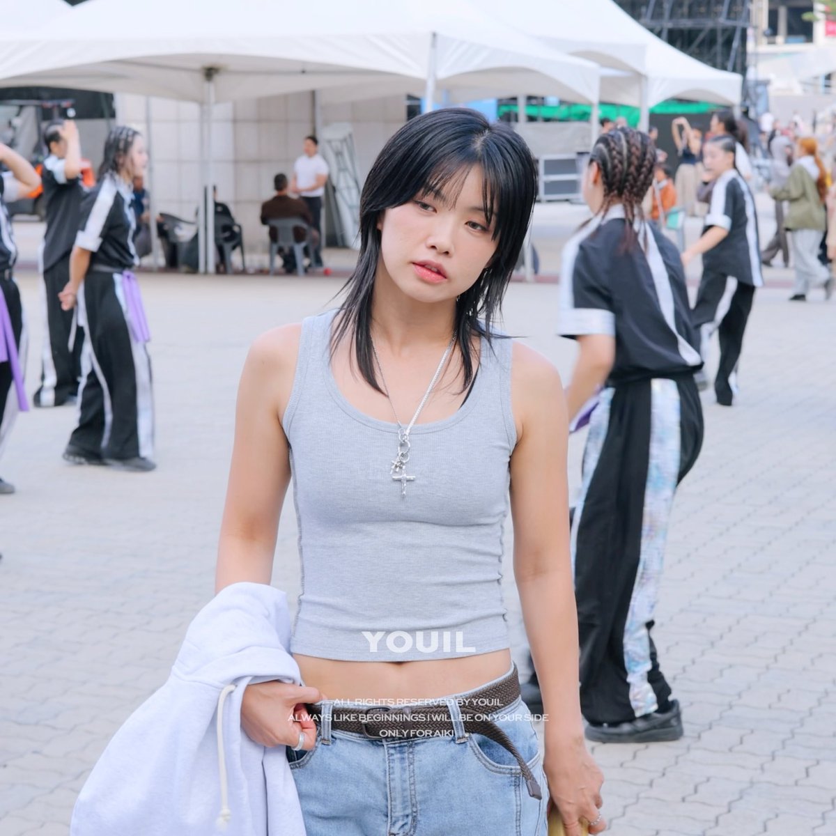 A young woman with dark hair wearing a grey tank top with 'YOUIL' branding stands at what appears to be a dance or performance event, with people in coordinated black outfits visible in the background.