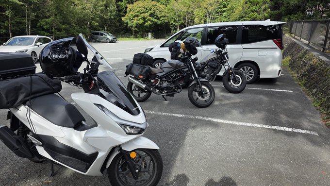 Two motorcycles, including a white scooter and a black bike with saddlebags, parked next to a white minivan in a parking lot.