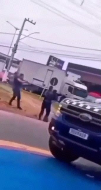A blurry scene shows armed individuals in tactical gear on a street near a white truck and a black police vehicle.