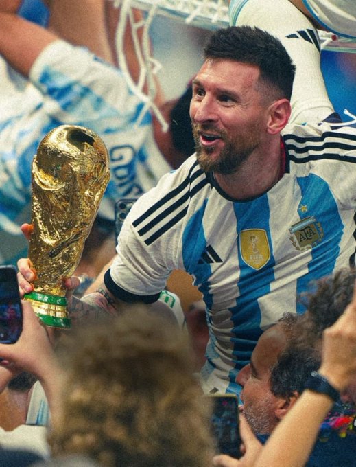 Lionel Messi wearing an Argentina national team jersey (light blue and white stripes) triumphantly holding the FIFA World Cup trophy while celebrating among a crowd.