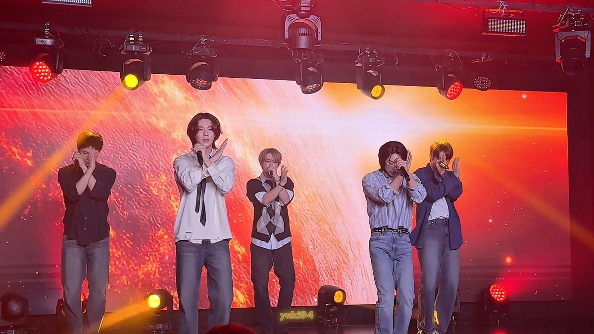 Five male performers in semi-formal attire sing and pose on stage with their arms crossed in an X gesture, set against a vibrant red and orange LED backdrop.