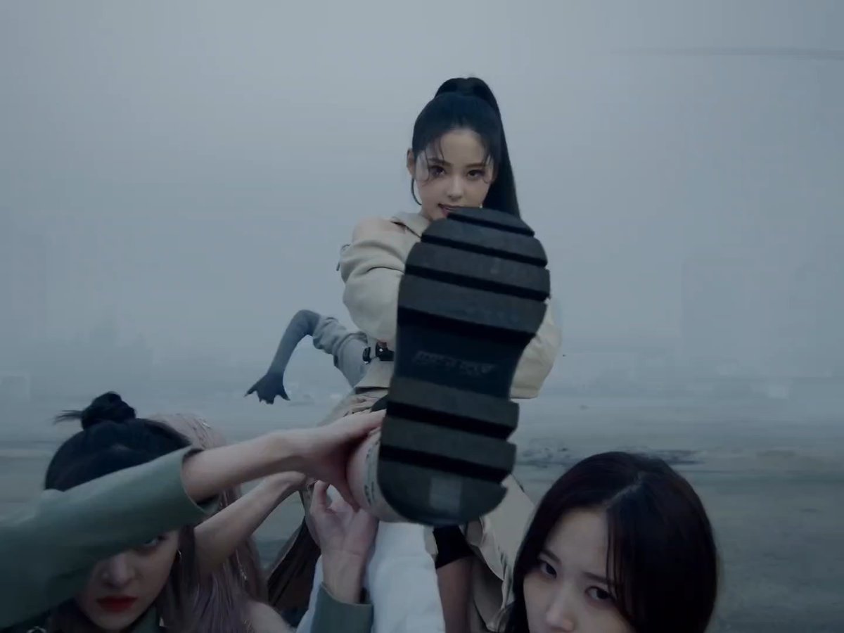 Several young women in beige and green outfits strike a dramatic pose outdoors in a misty environment, with one prominently displaying the sole of her sneaker toward the camera.