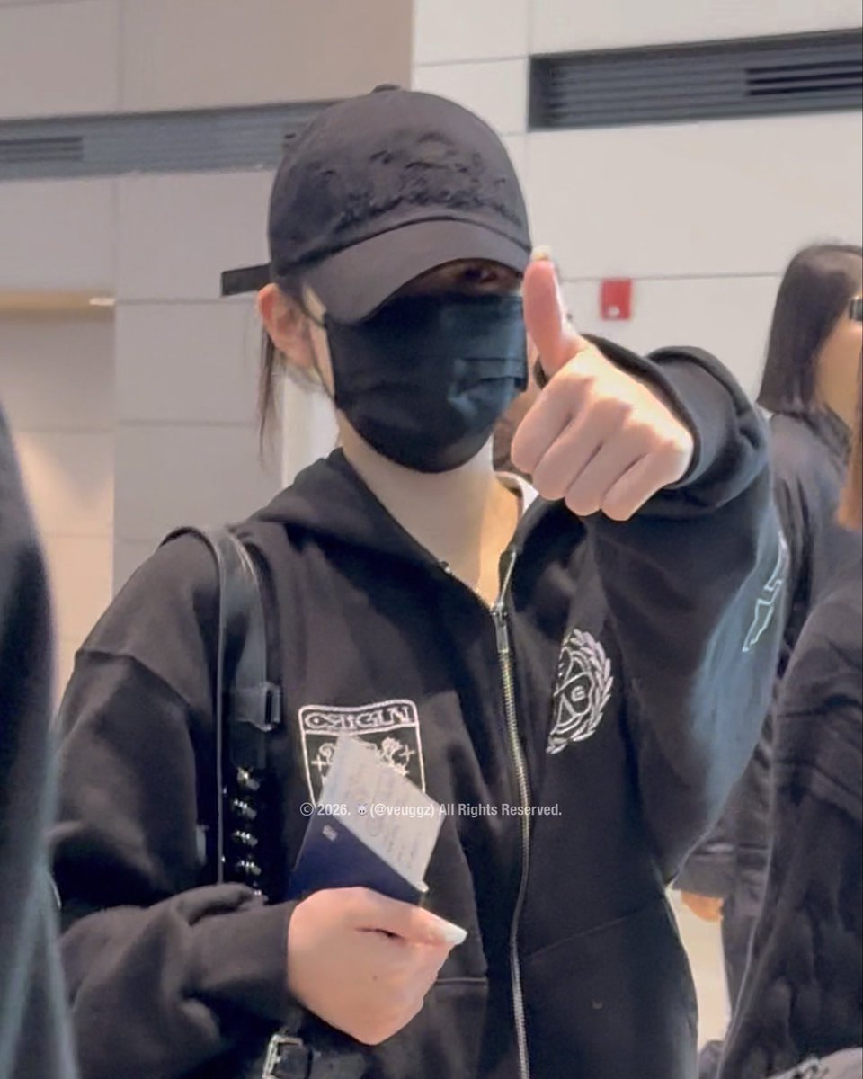A woman wearing a black cap with embroidered designs, a black face mask, and a black zip-up jacket giving a thumbs-up gesture while holding a passport at an airport.