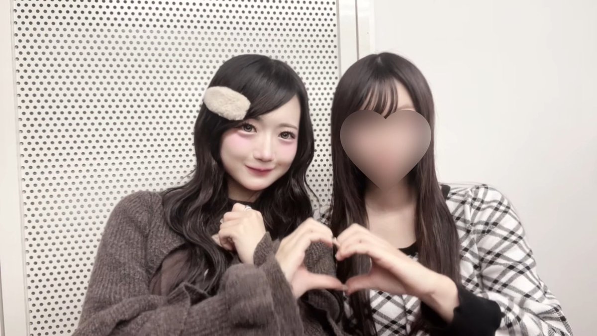 Two women pose together against a perforated background; the one on the right has a heart emoji covering her face, while both form a heart shape with their hands.