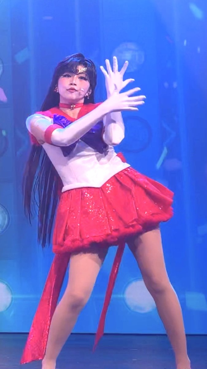A photo of a woman performing on stage in a red and white Sailor Moon-style costume with a sparkly red skirt, posed with her hands extended forward against a blue-lit background.