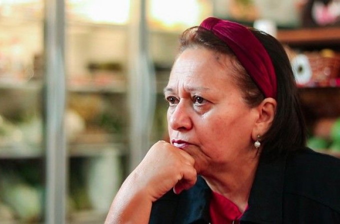 Fátima Bezerra, the former governor of Rio Grande do Norte and current senator, sits thoughtfully with her chin resting on her hand, wearing a red headband and pearl earrings. The image aligns with the post celebrating her political career and role in expanding the PT's presence in the state.