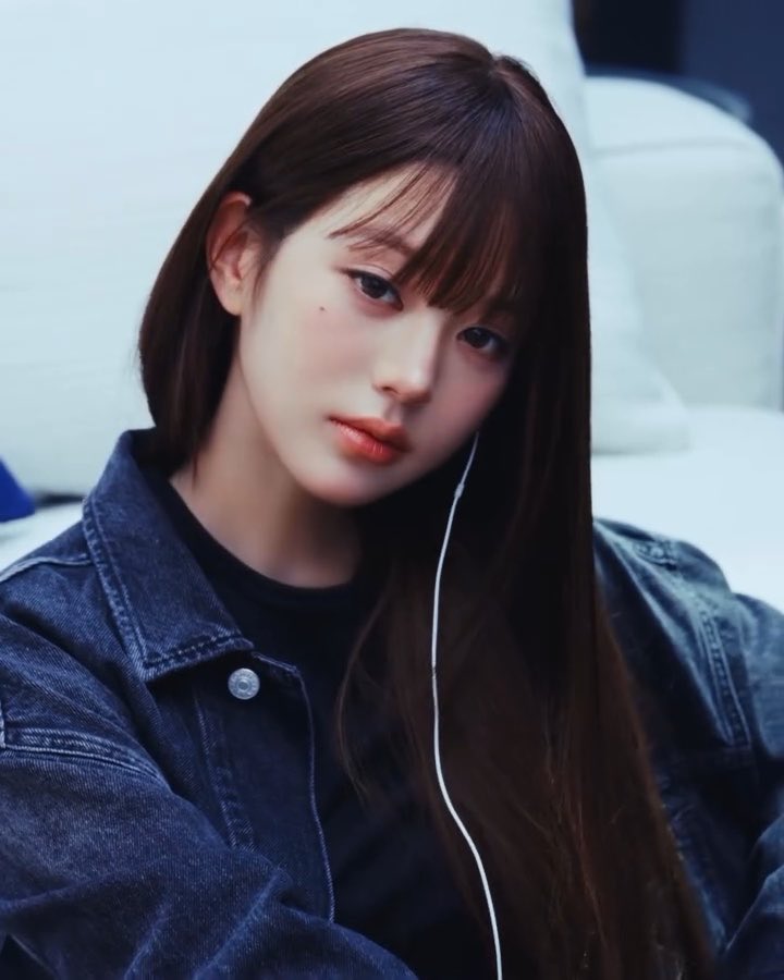 A portrait of a young woman with long dark hair and bangs, wearing a denim jacket and earphones, looking directly at the camera.