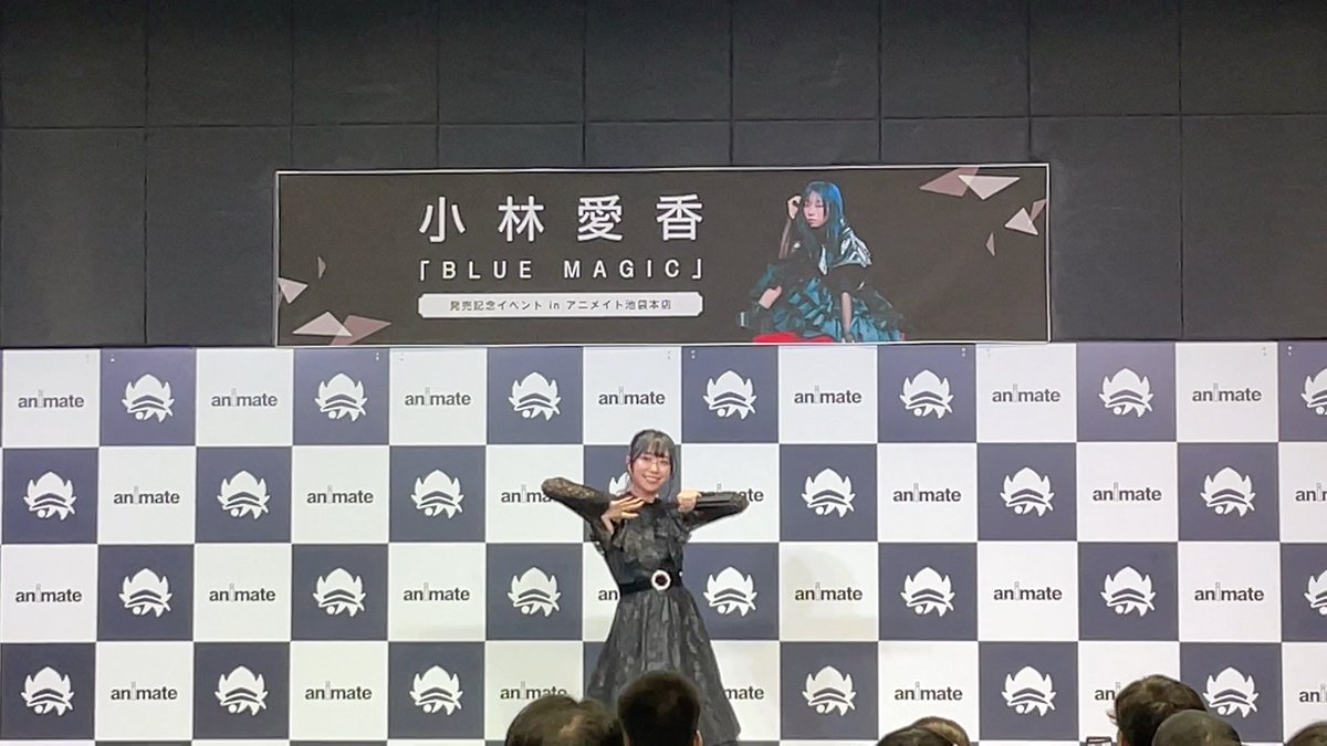 An idol in a black dress poses with both hands under their chin at an Animate event, with a banner above reading '小林愛香 「BLUE MAGIC」' (Aiko Kobayashi - BLUE MAGIC).