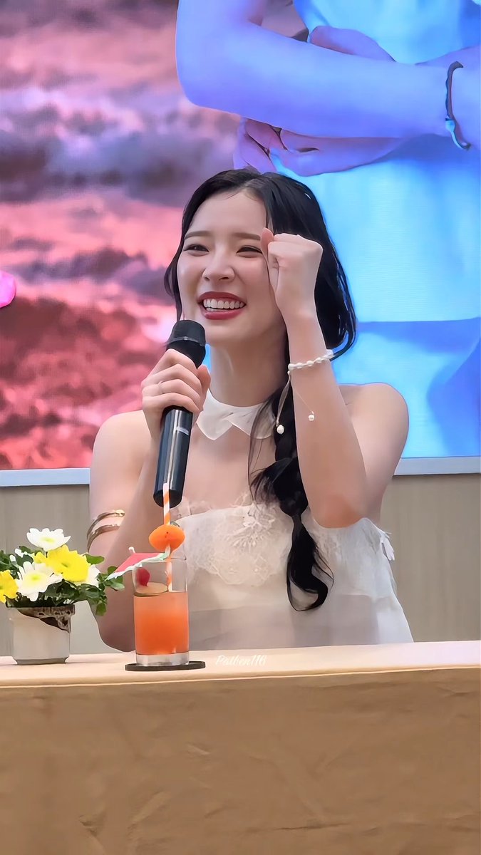 A smiling woman with dark hair holds a microphone while wearing a white dress, with a drink and flowers on the table in front of her.
