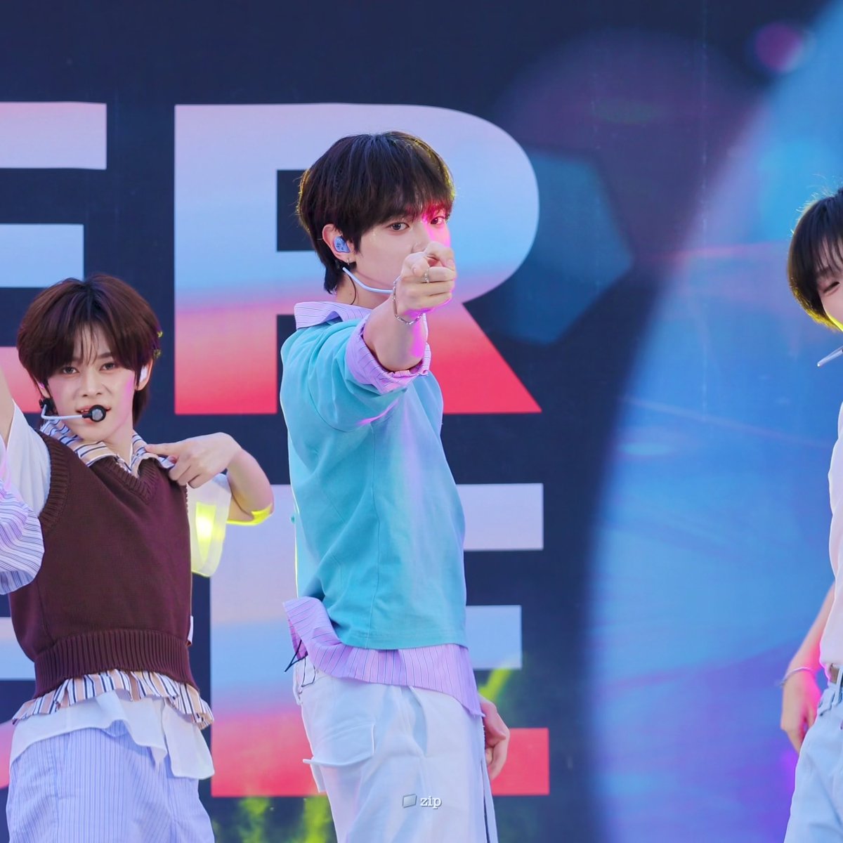 A young male K-pop performer in a light blue sweater pointing directly at the camera during a stage performance, with another performer visible beside him.