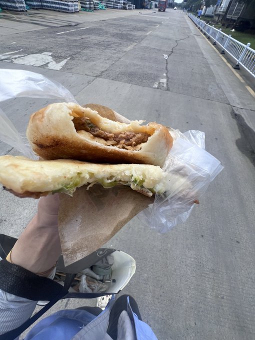 A hand holds a partially eaten stuffed flatbread sandwich wrapped in paper and plastic, with a street and sidewalk visible in the background.