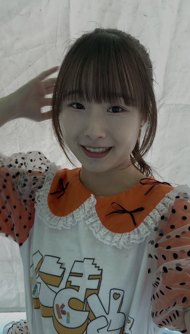 A smiling young woman with brown bangs wears a white t-shirt with orange polka dot sleeves and an orange collar, posing indoors against a white sheet.