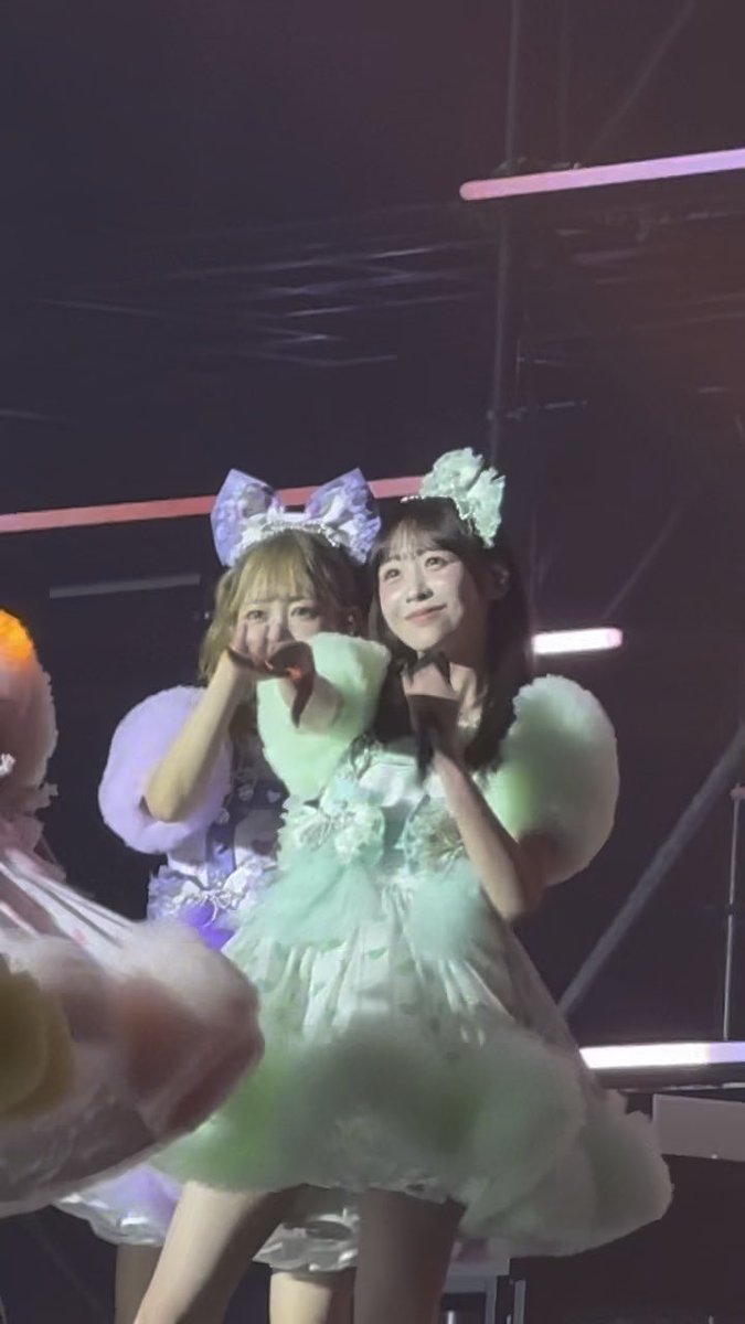 Two performers in elaborate green and lavender frilly costumes with bow headpieces pose together on a dimly lit stage, making heart gestures with their hands.