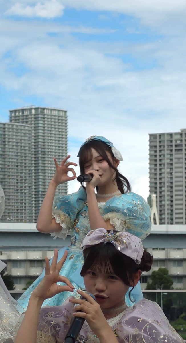 Two young women in ornate blue and lavender dresses pose with microphones and make 'OK' signs, set against a backdrop of tall buildings and a blue sky.