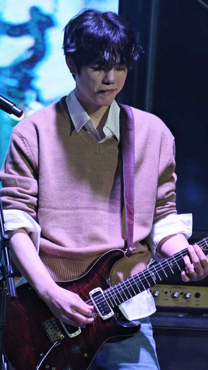 A young man with dark hair wearing a tan sweater over a white collared shirt, playing a red electric guitar on stage.