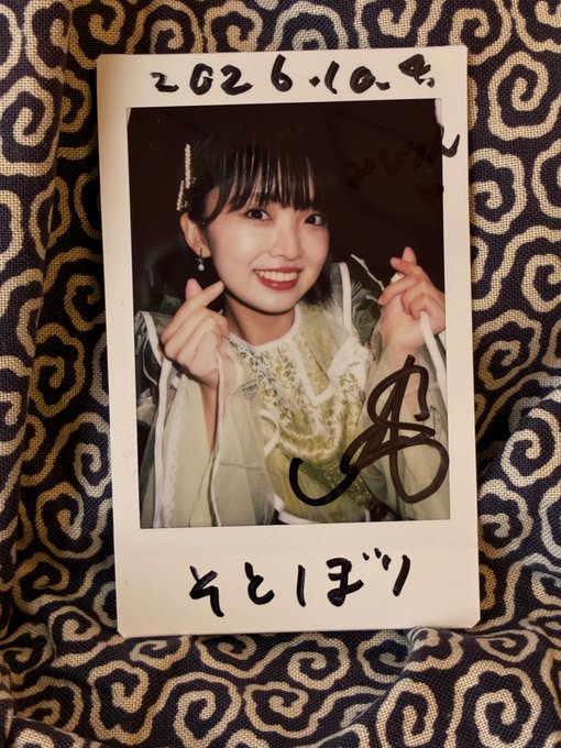 A polaroid photo of a smiling young woman making finger heart gestures, with handwritten Japanese text reading "2026.10.4" and "そとはづり" and a signature on it.