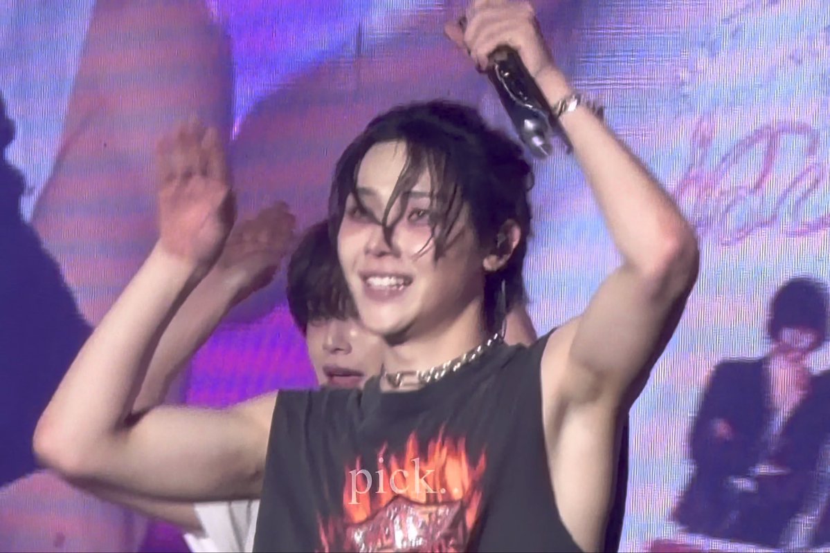 A concert photo of a young performer smiling while holding a microphone, wearing a black flame-patterned sleeveless shirt with 'pick.' text and a silver chain necklace.