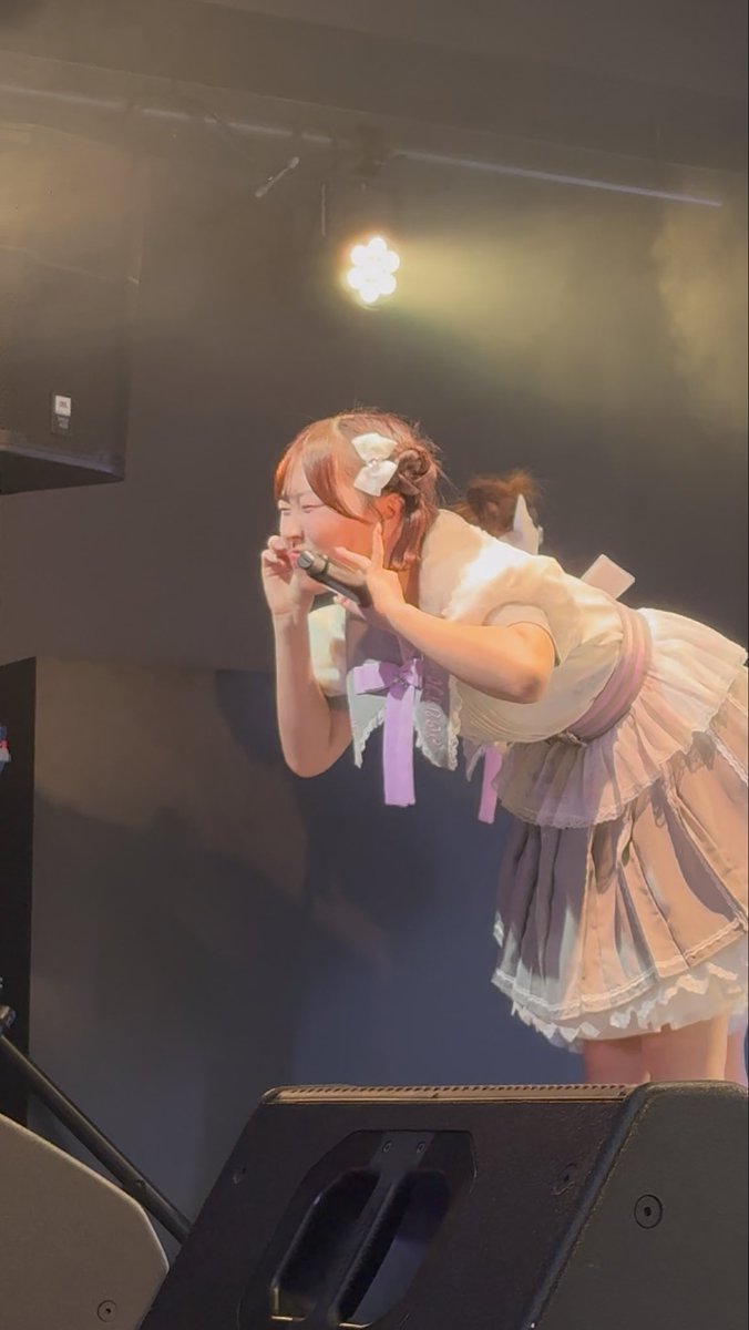A young woman in a frilly white and lavender maid-style dress with a hair bow leans forward on stage, singing passionately into a microphone under a spotlight with stage speakers visible in the foreground.