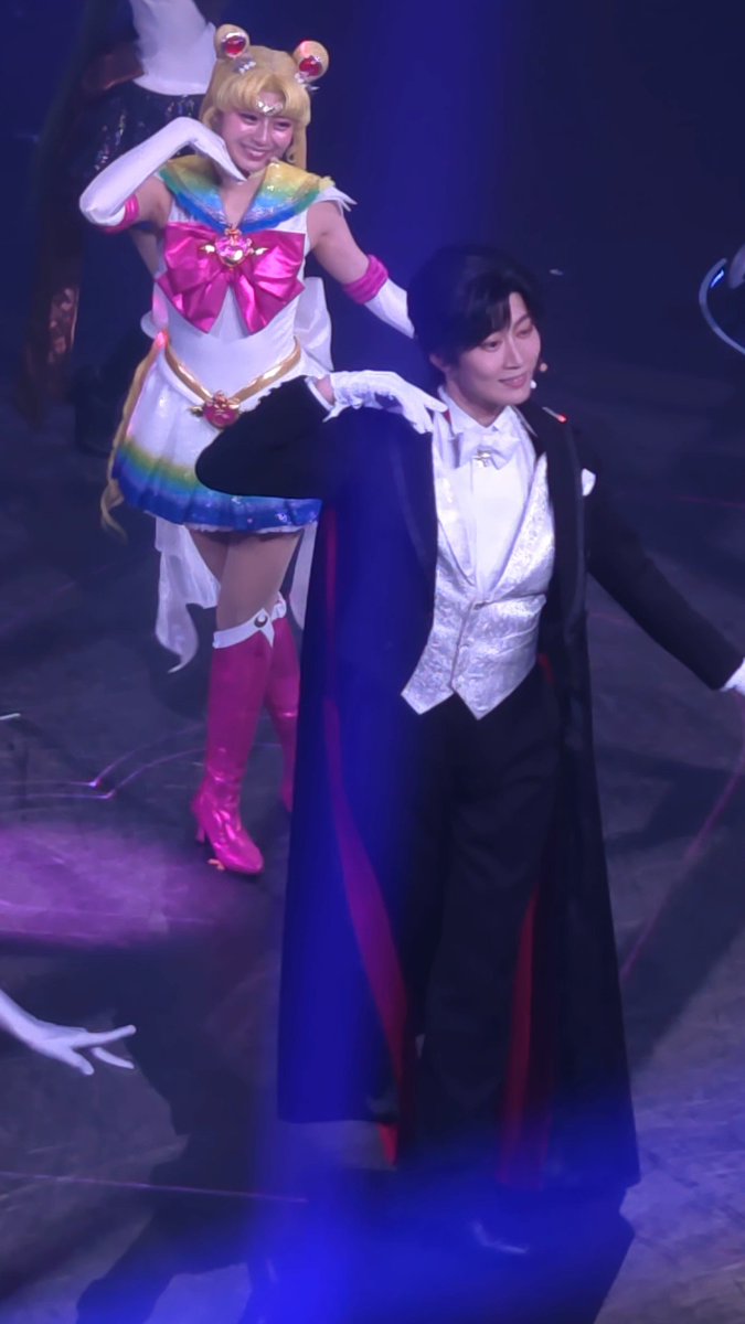 Two performers on stage in costume; one dressed as Super Sailor Moon with blonde hair and a colorful outfit, and the other in a formal tuxedo with a cape.