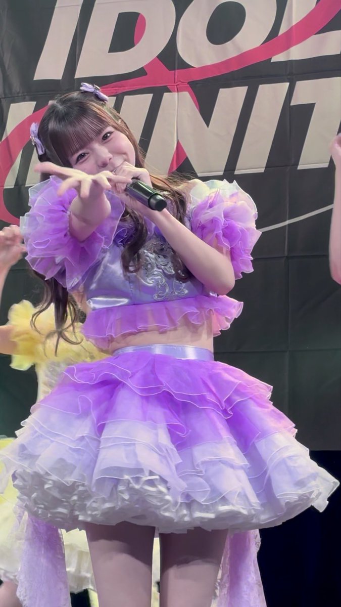 Live concert photo of a young female idol performing in a voluminous purple ruffled two-piece outfit with matching hair accessories, holding a microphone and pointing toward the camera, with 'IDOL' branding visible on the backdrop