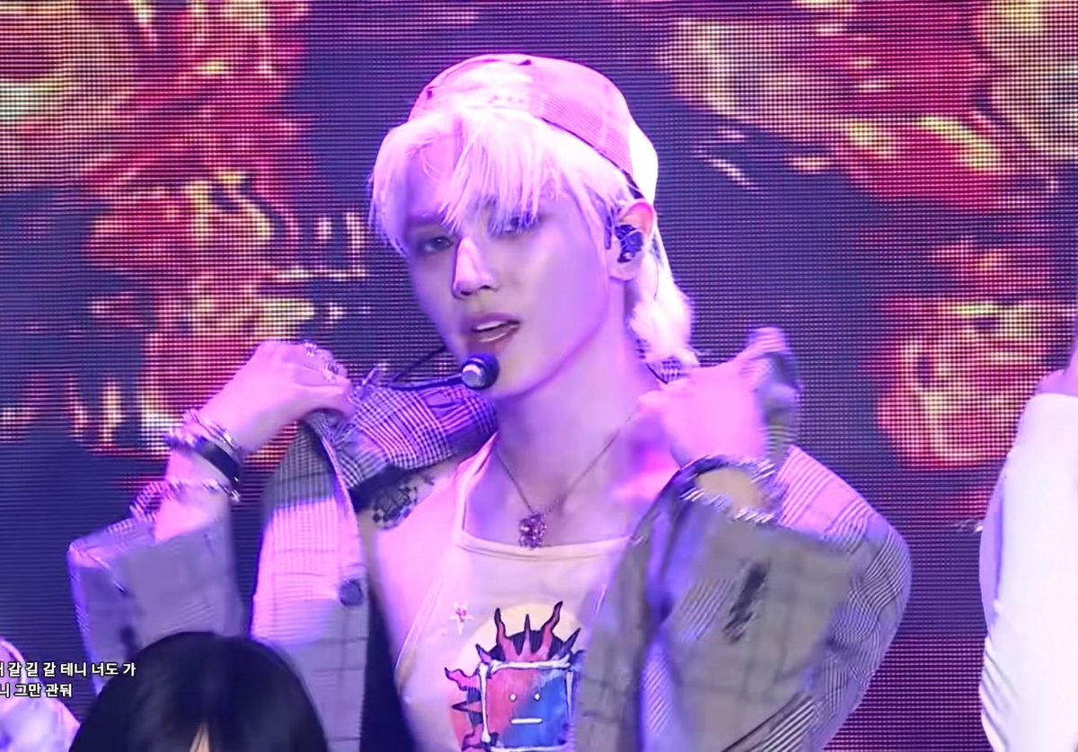 A performer with light pink/blonde hair and a backward cap on stage, wearing a graphic tank top and holding a microphone, with Korean text in the bottom left corner.