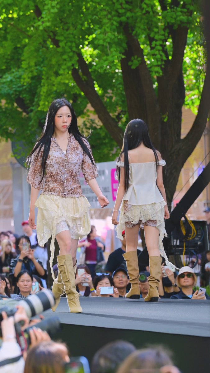 Two young female models walk on a runway during a fashion show, wearing sheer layered outfits and tan knee-high boots, with an audience photographing them.